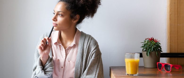 Woman thinks as she sits next to a table with orange juice, a plant, and sunglasses.