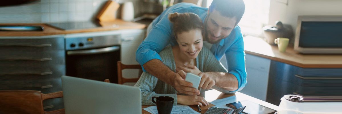Couple looking at a mobile phone while reviewing paperwork in their kitchen