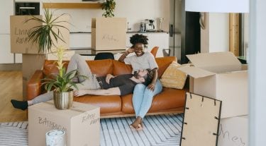 A couple relaxes on a couch surrounded by unpacked moving boxes in their living room.