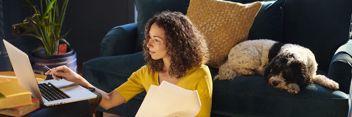 A woman works on financial paperwork at her laptop while her dog sleeps on the sofa behind her.