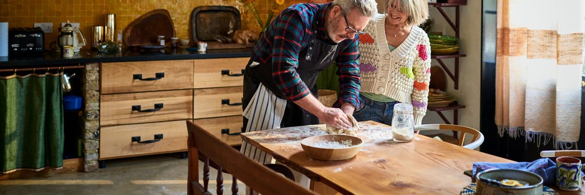 A retired couple roll out bread dough together on their dining table.
